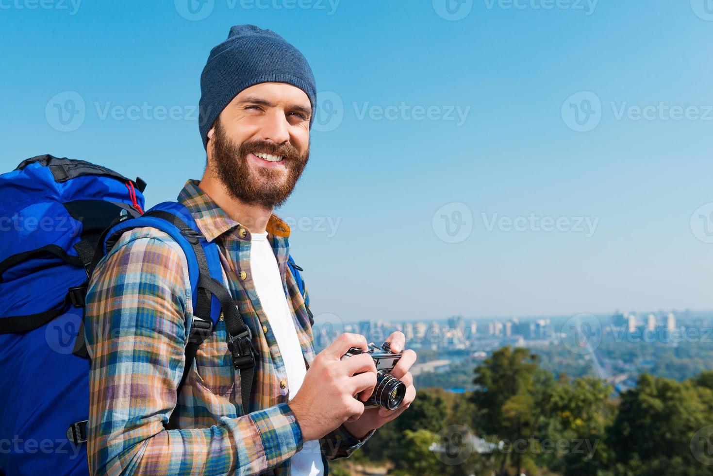 enregistrer chaque vue en mémoire. beau jeune homme portant un sac à dos et regardant la caméra à travers les épaules avec le sourire tout en se tenant dans la nature et en tenant la caméra photo