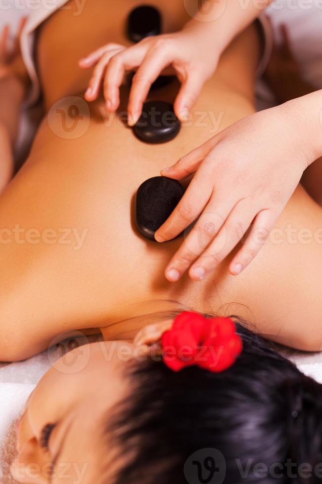 massage aux pierres chaudes. vue de dessus de la belle jeune femme allongée sur le devant pendant que le massothérapeute lui masse le dos avec des pierres de spa photo