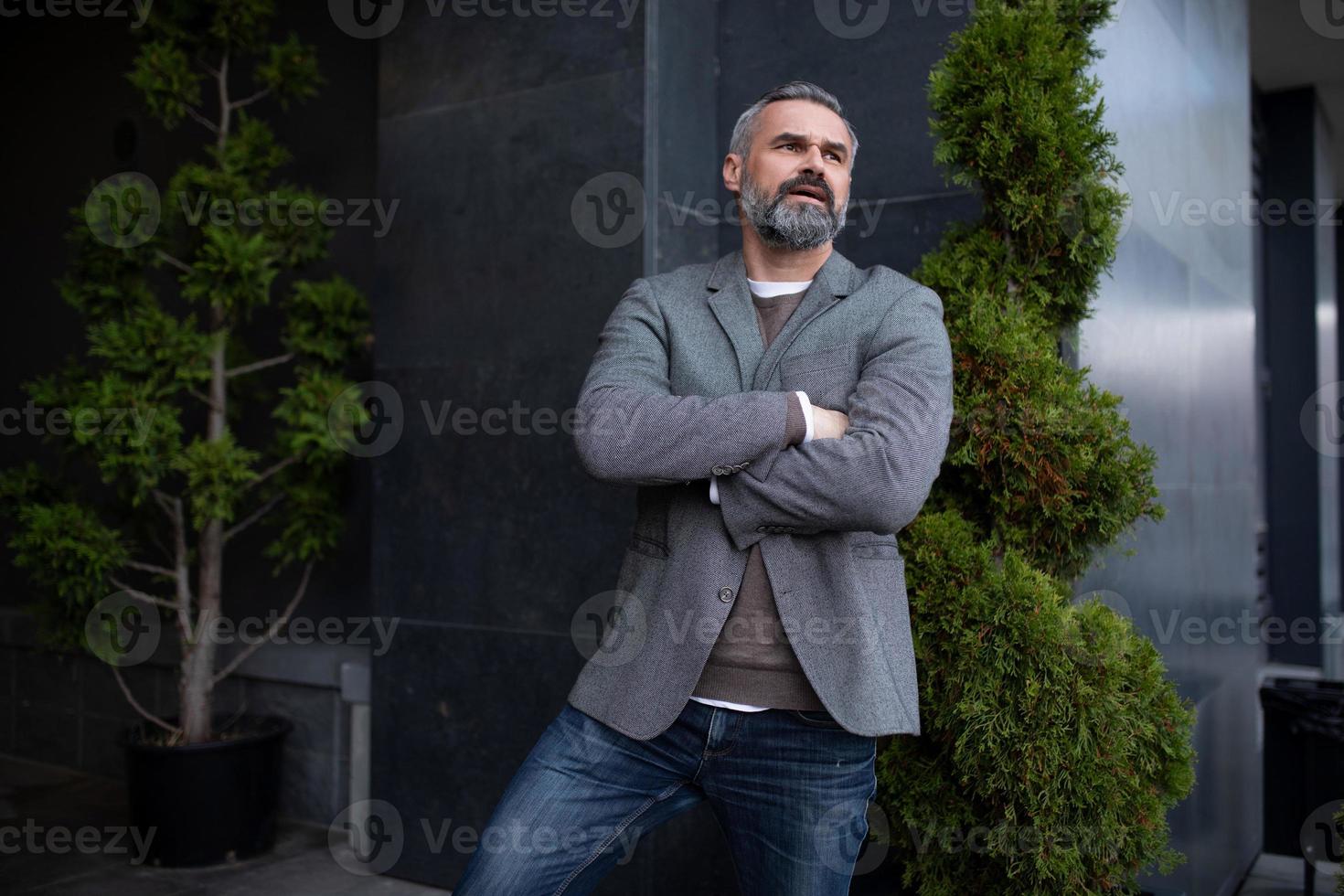 un homme d'affaires âgé sérieux dans une veste grise à côté de l'entrée de l'immeuble de bureaux regarde au loin dans la pensée photo