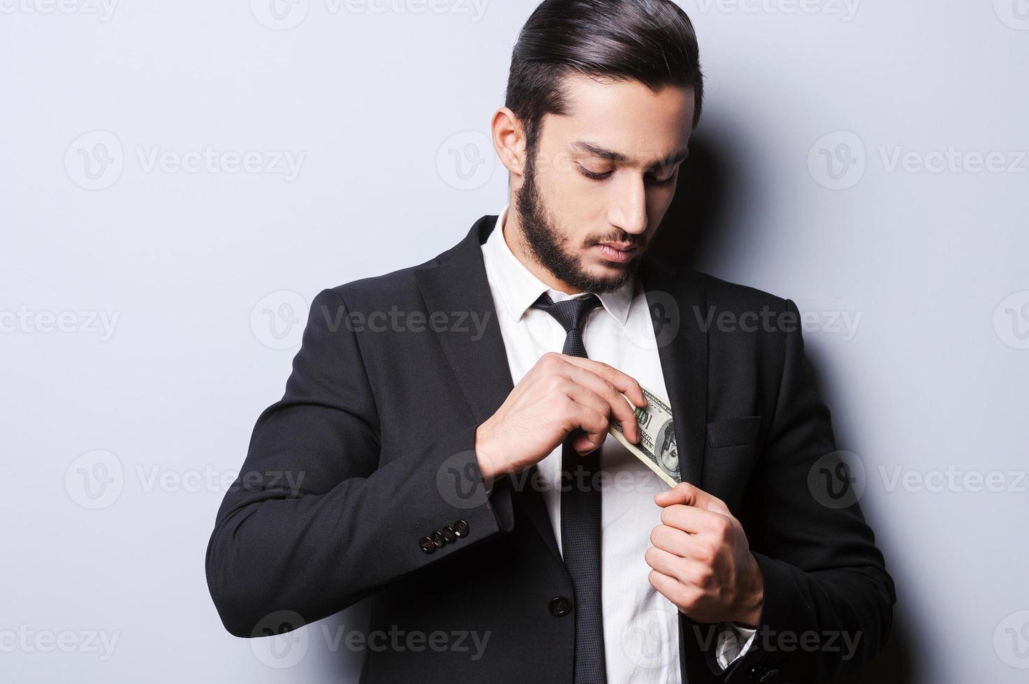 j'aime l'argent et ils m'aiment. beau jeune homme en tenues de soirée mettant de l'argent dans sa poche en se tenant debout sur fond gris photo