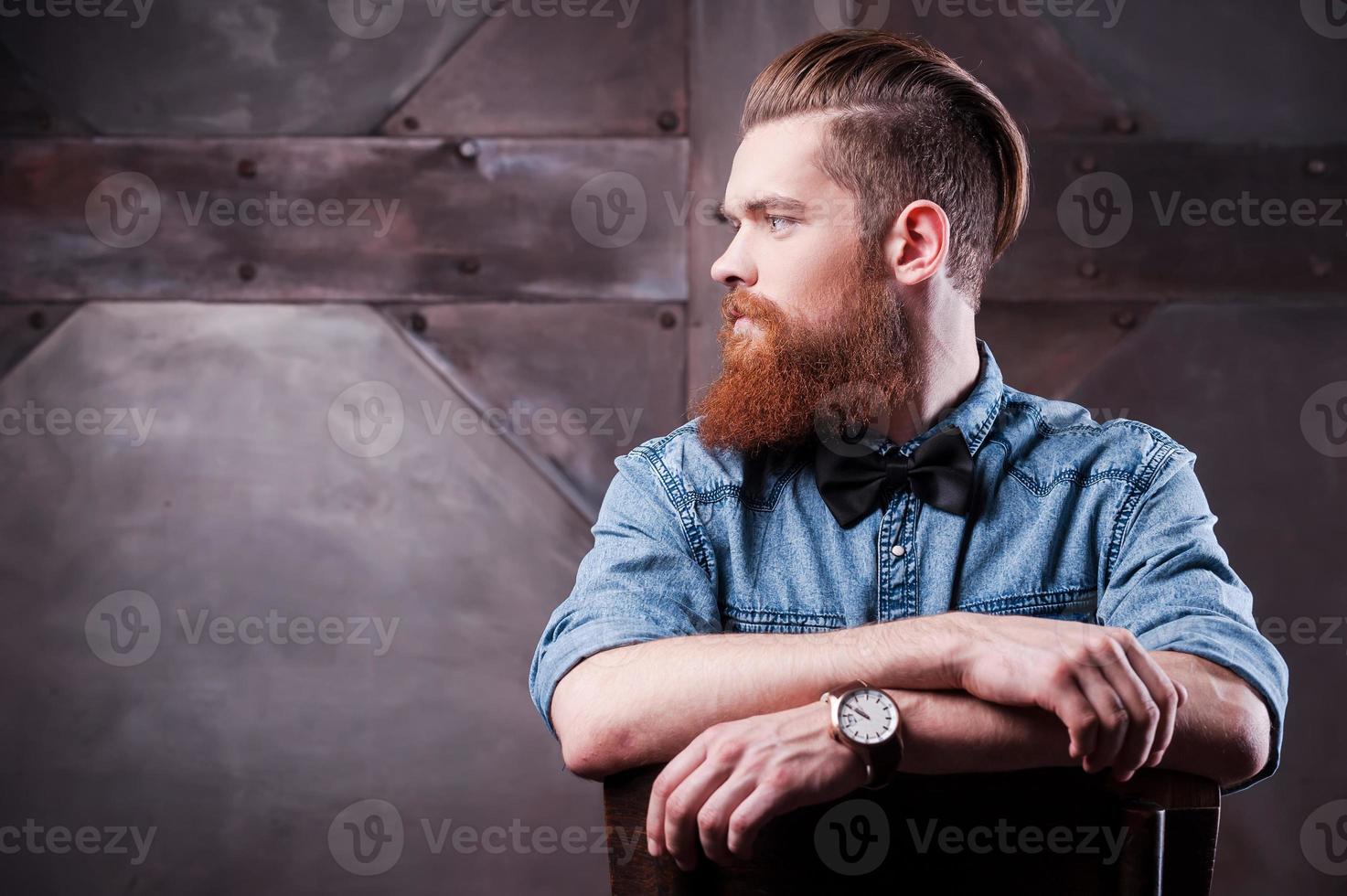 confiant dans son style parfait. profil d'un beau jeune homme barbu regardant loin et assis sur une chaise photo