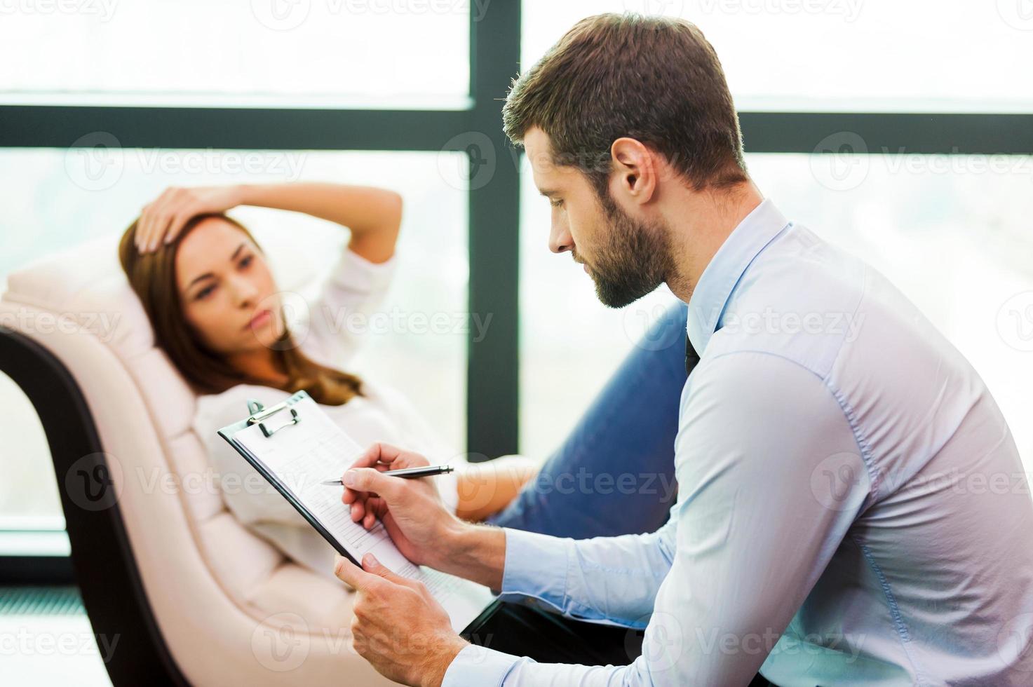 elle a besoin d'un avis d'expert. jeune femme frustrée assise à la chaise et tenant la main sur la tête tandis que le jeune homme assis près d'elle et écrivant quelque chose dans son presse-papiers photo