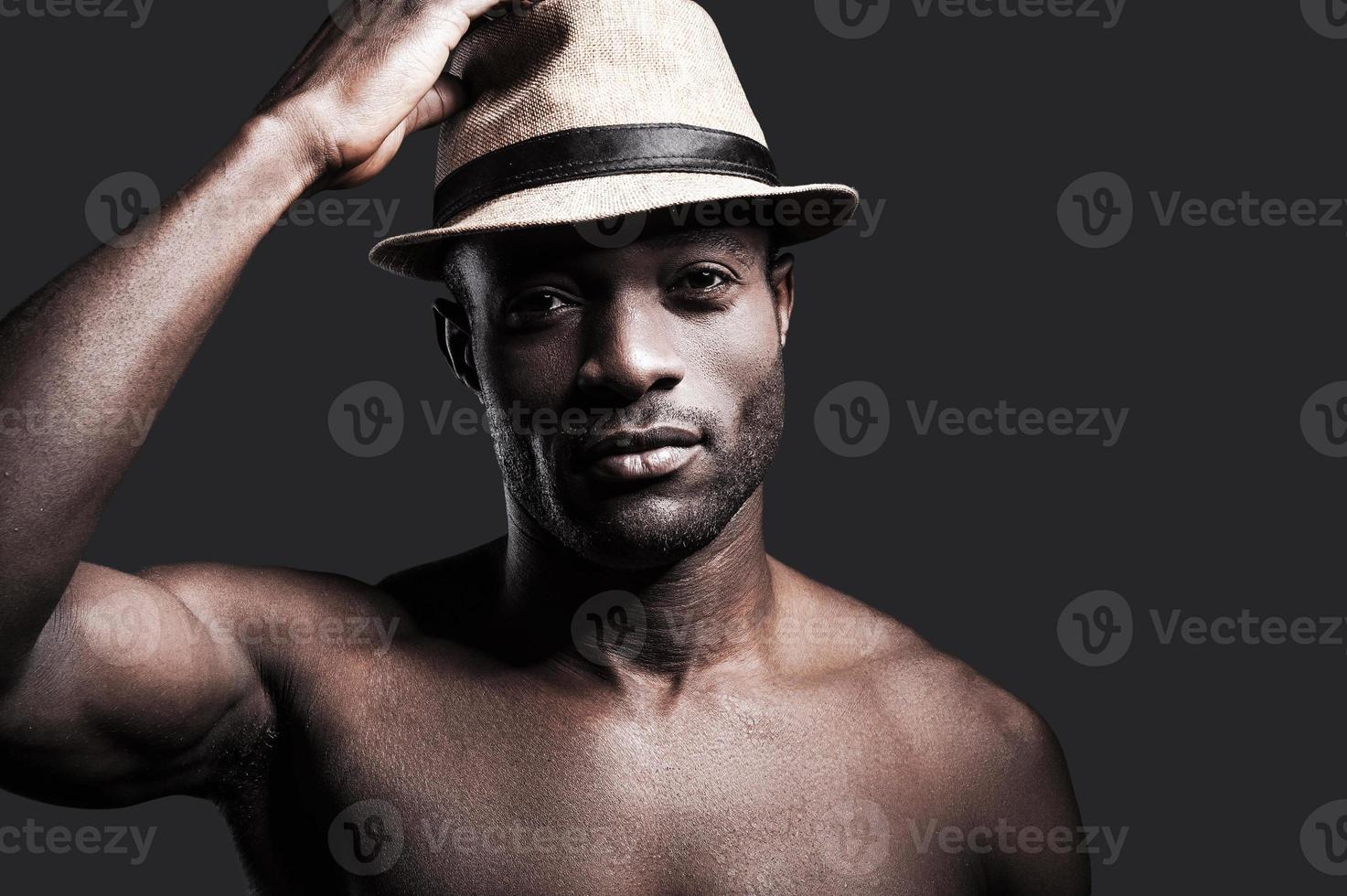 mon chapeau porte-bonheur. portrait de jeune homme africain torse nu ajustant son chapeau et regardant la caméra en se tenant debout sur fond gris photo