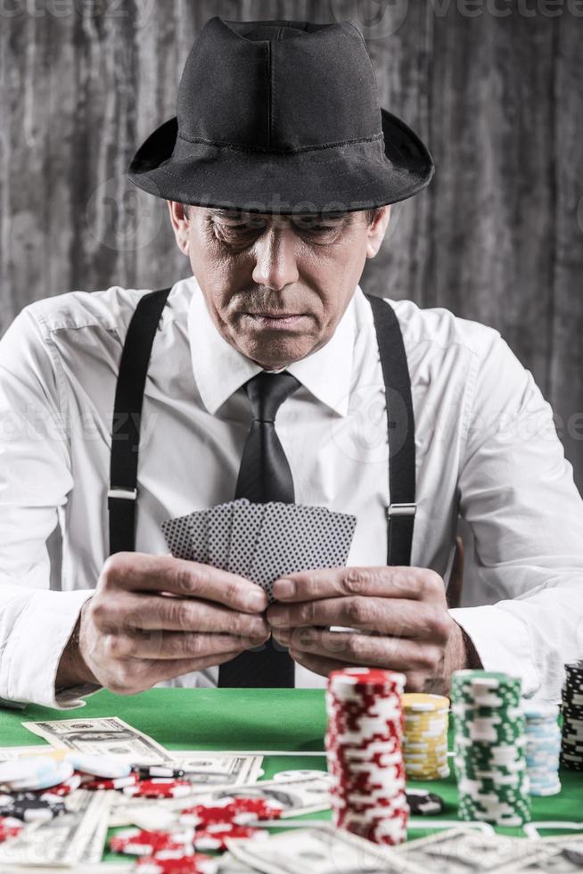 joueur de poker confiant. gros plan d'un homme senior sérieux en chemise et bretelles assis à la table de poker et tenant des cartes avec de l'argent et des jetons de jeu posés tout autour de lui photo