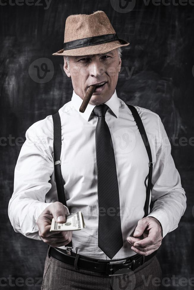 l'argent n'est pas un problème. homme âgé sérieux en chapeau et bretelles fumant un cigare et tendant de l'argent en se tenant debout sur un fond sombre photo