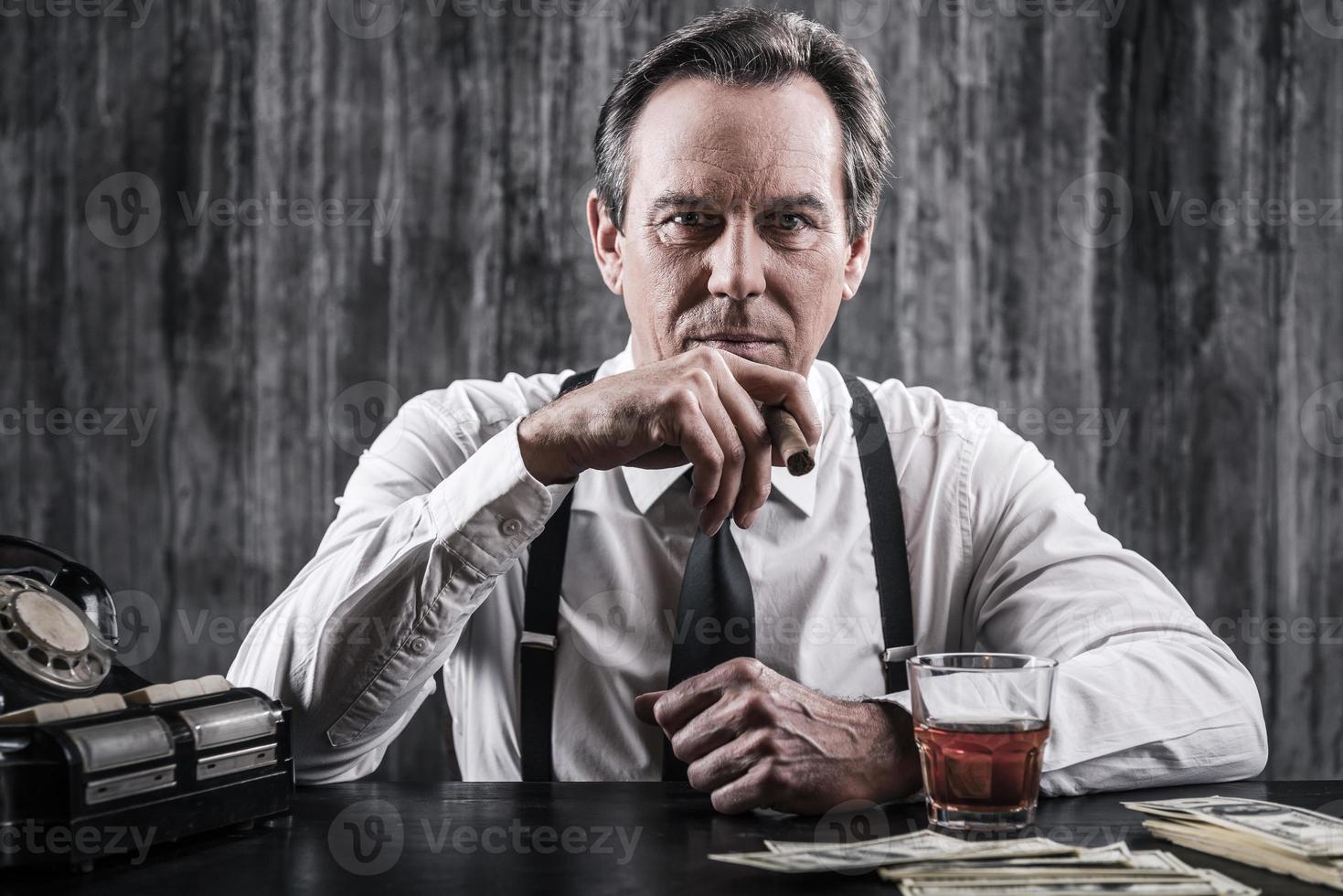 patron de la mafia. homme âgé sérieux en chemise et bretelles assis à la table et fumant un cigare tandis que beaucoup d'argent se trouvait près de lui photo