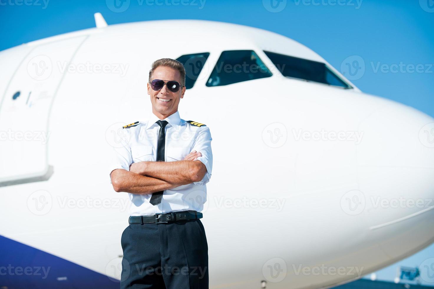 le ciel est ma passion. pilote masculin confiant en uniforme gardant les bras croisés et souriant avec un avion en arrière-plan photo