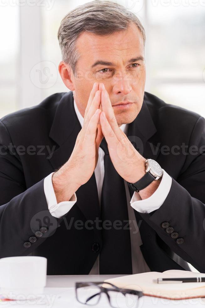 perdu dans ses pensées professionnelles. homme mûr réfléchi en tenues de soirée tenant les mains jointes et regardant loin tout en étant assis sur son lieu de travail photo