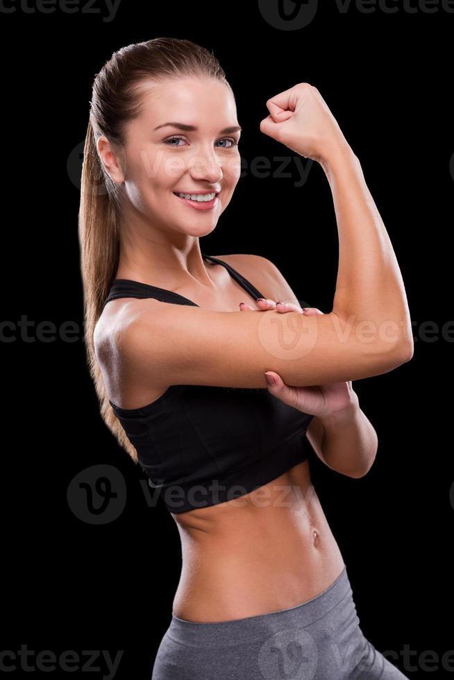 mesurer son biceps parfait. jeune femme sportive touchant son biceps et souriant debout sur fond noir photo