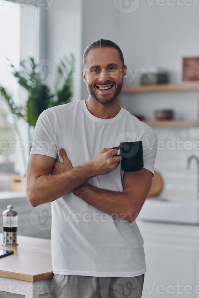 jeune homme appréciant le café et souriant tout en se tenant à la cuisine photo