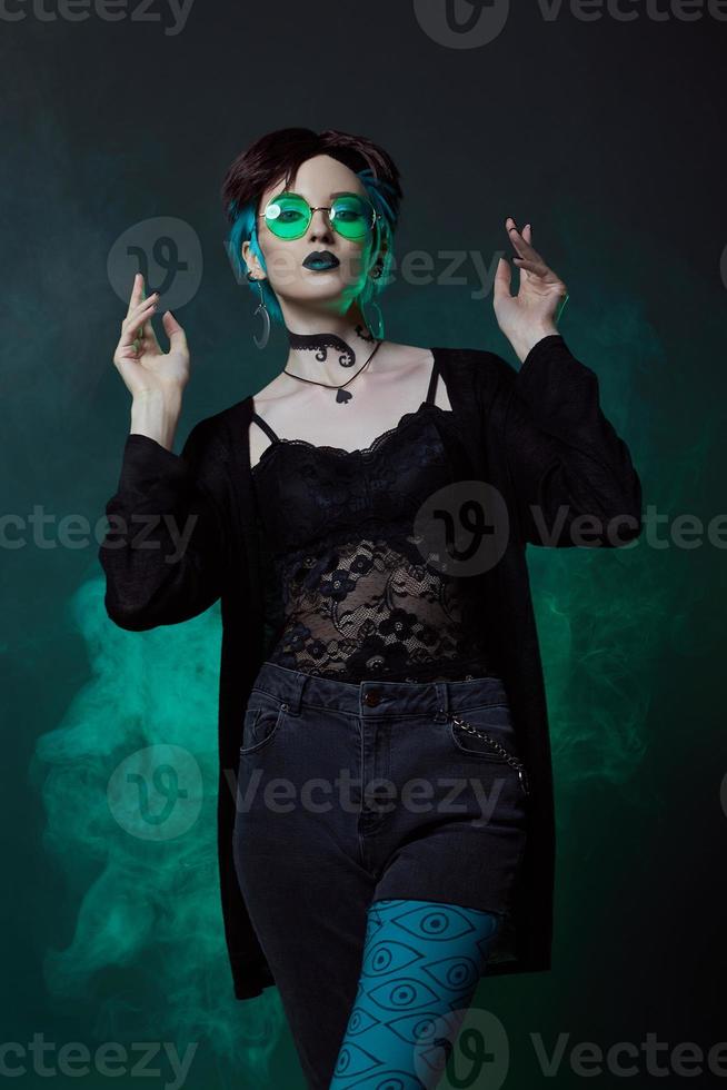 jeune sorcière d'halloween dans un chapeau rond et des lunettes rondes vertes sur fond vert foncé. cheveux teints, rouge à lèvres foncé, boucles d'oreilles croissant de lune photo