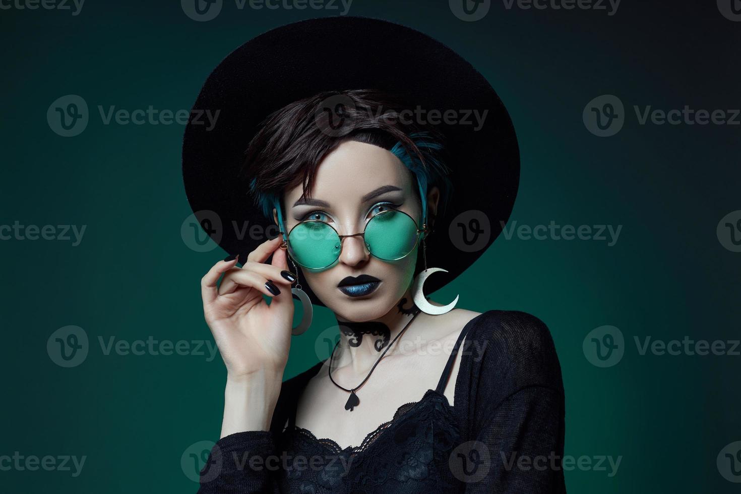 jeune sorcière d'halloween dans un chapeau rond et des lunettes rondes vertes sur fond vert foncé. cheveux teints, rouge à lèvres foncé, boucles d'oreilles croissant de lune photo