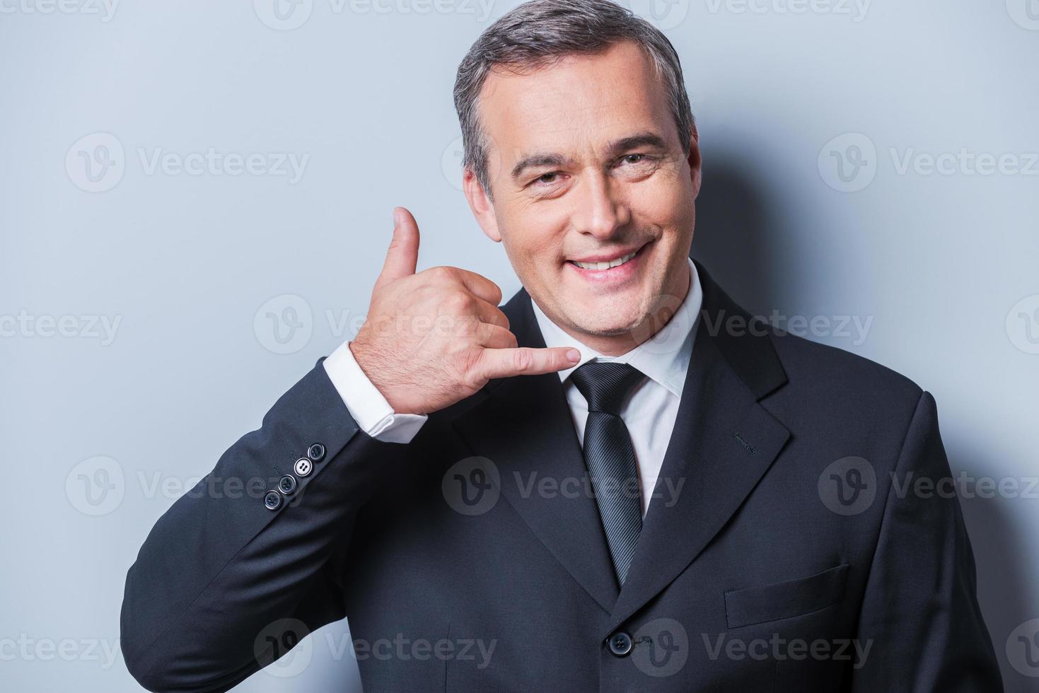 en attente de votre appel. homme mûr confiant en tenues de soirée gesticulant téléphone portable près de son visage et souriant en se tenant debout sur fond gris photo