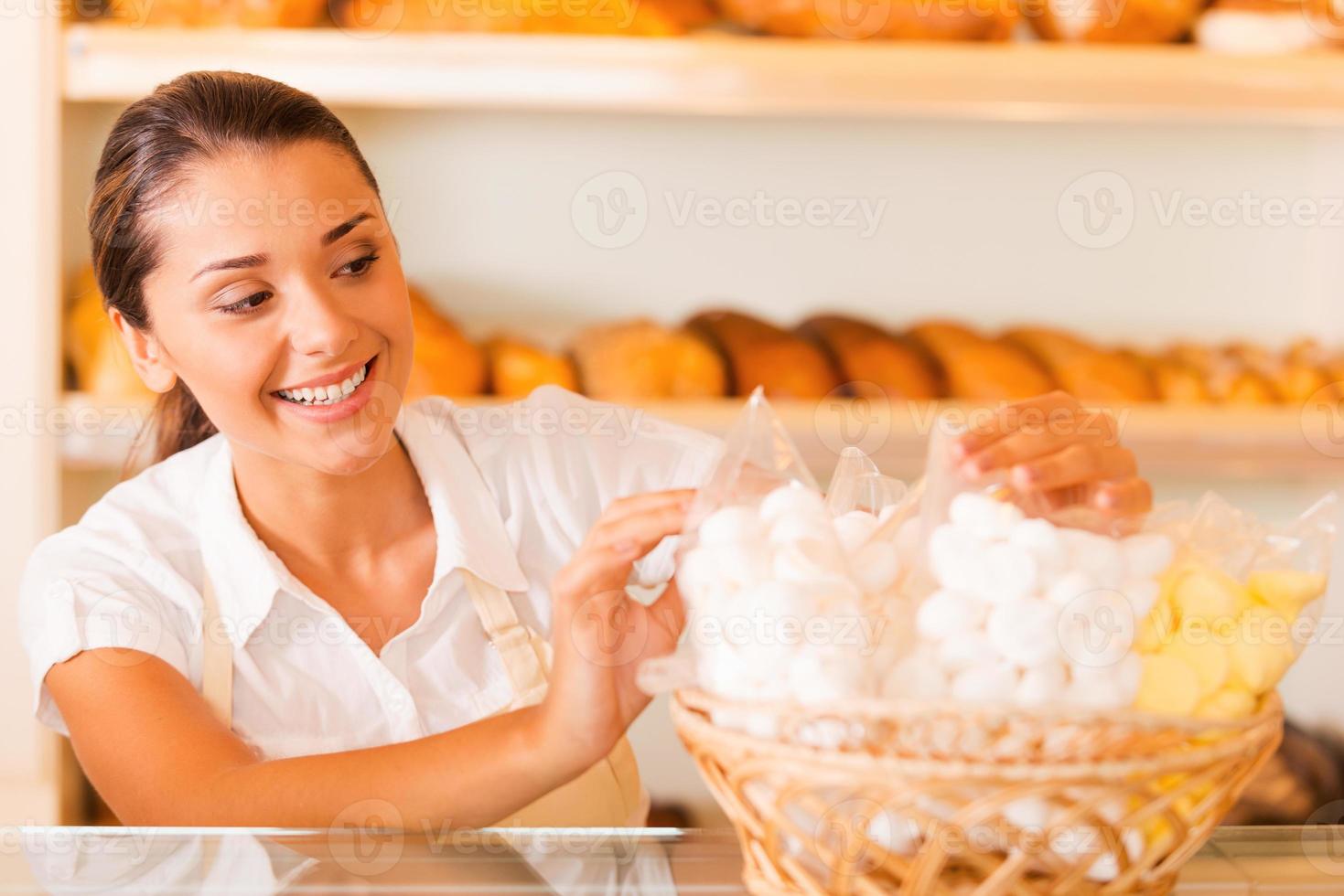 emballer des biscuits frais à vendre. belle jeune femme en tablier emballant des biscuits et souriant debout dans une boulangerie photo