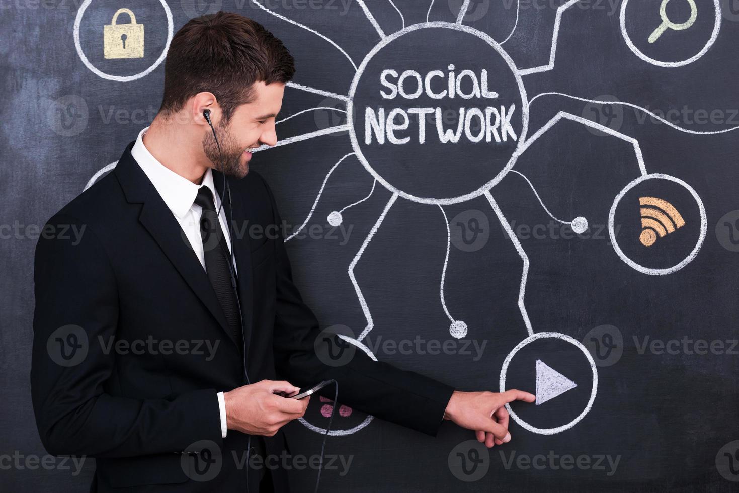 belle fonction pour se détendre. beau jeune homme au casque tenant le téléphone en se tenant debout contre le dessin à la craie du réseau social sur le tableau noir photo