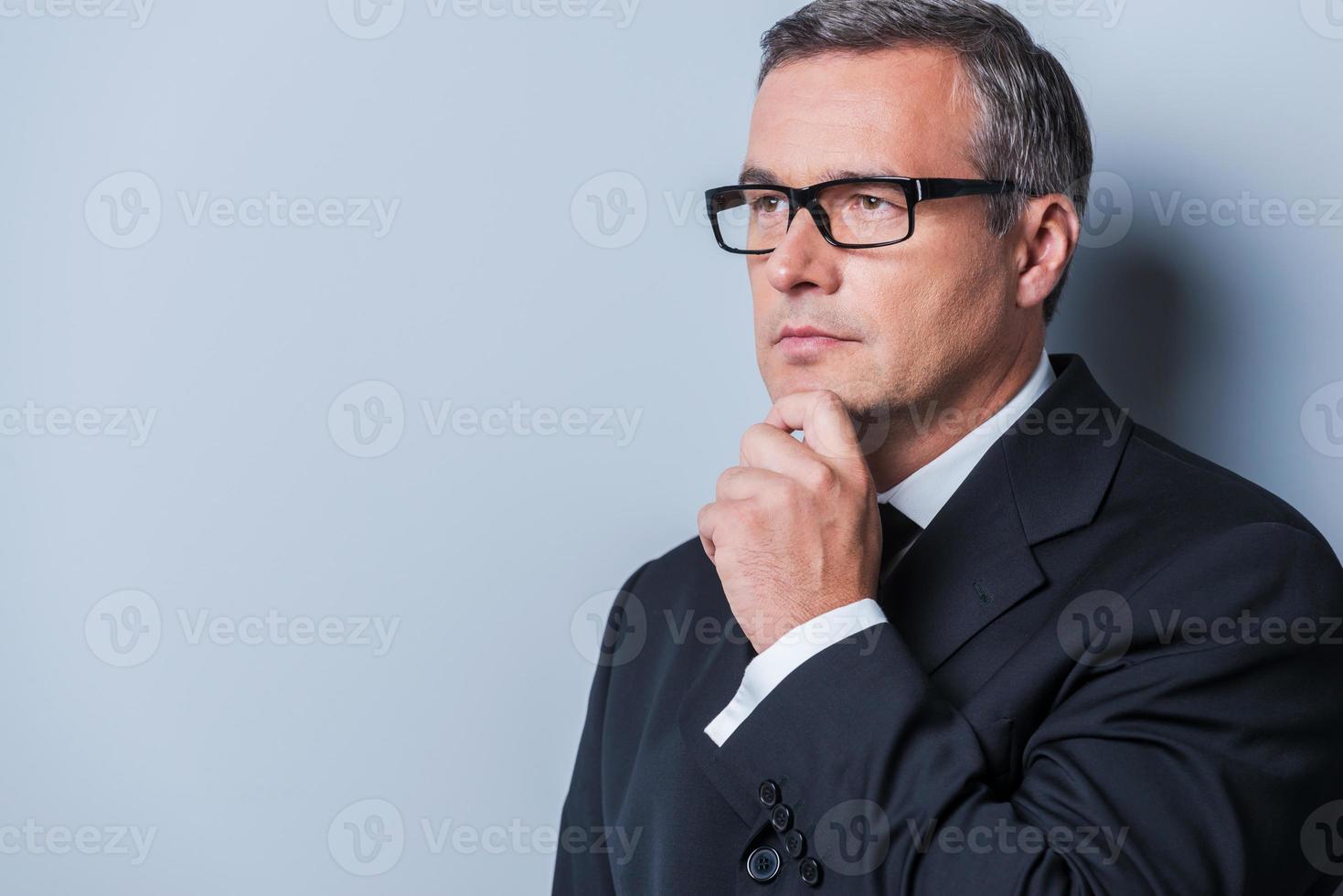 réfléchir à des solutions. homme d'âge mûr réfléchi en tenues de soirée tenant la main sur le menton et en détournant les yeux en se tenant debout sur fond gris photo