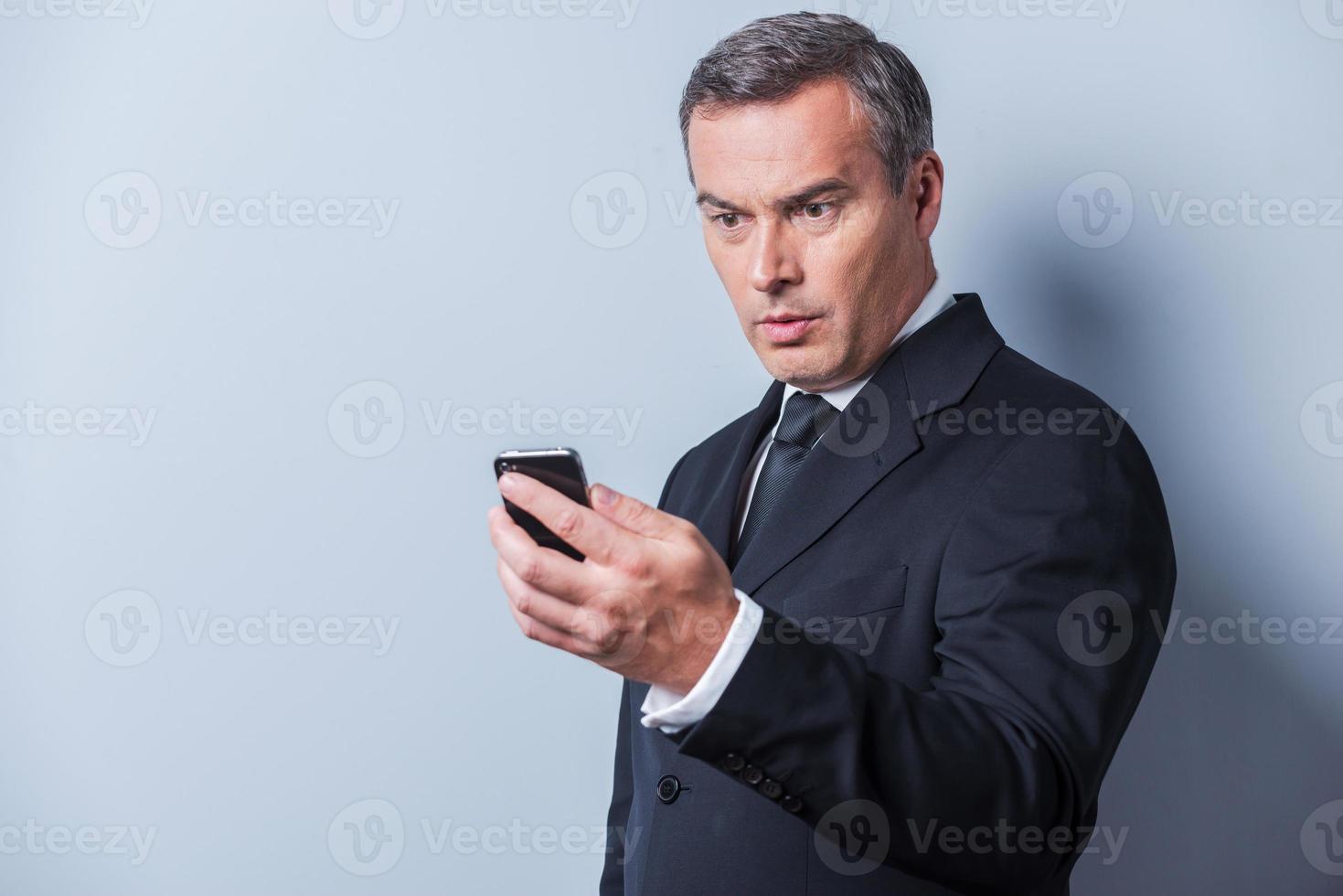 nouvelle choquante. homme mûr surpris en tenues de soirée tenant un téléphone portable et le regardant en se tenant debout sur fond gris photo