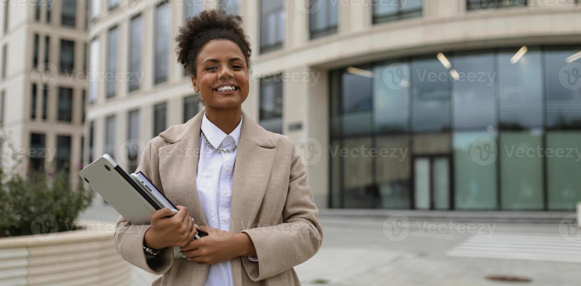 portrait d'une jeune femme afro-américaine réussie avec un ordinateur portable dans les mains et un large sourire sur fond d'immeuble de bureaux, concept de négociations commerciales réussies photo