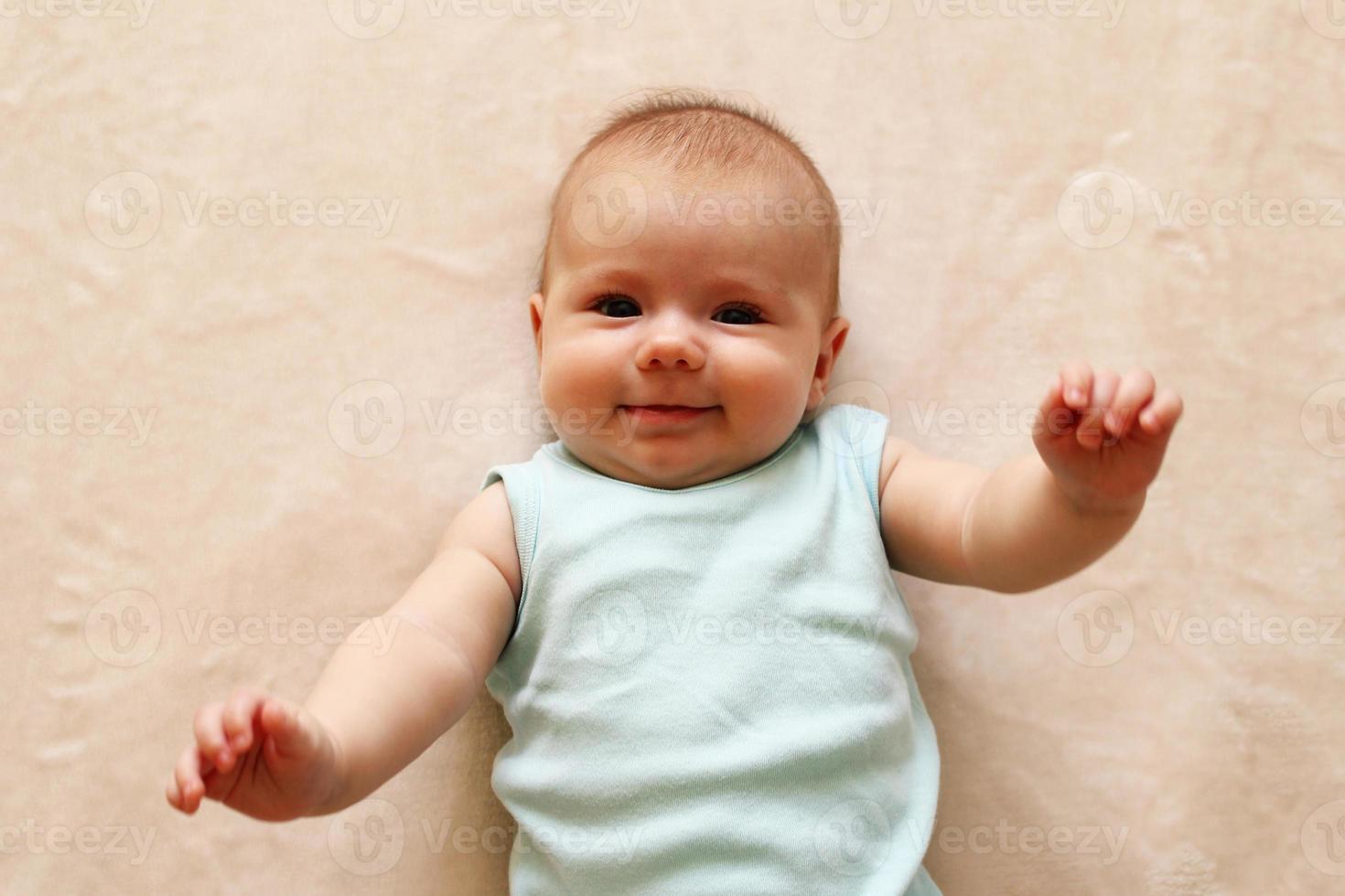 portrait d'un bébé souriant de trois mois en body bleu est allongé sur une couverture beige et regarde la caméra. photo