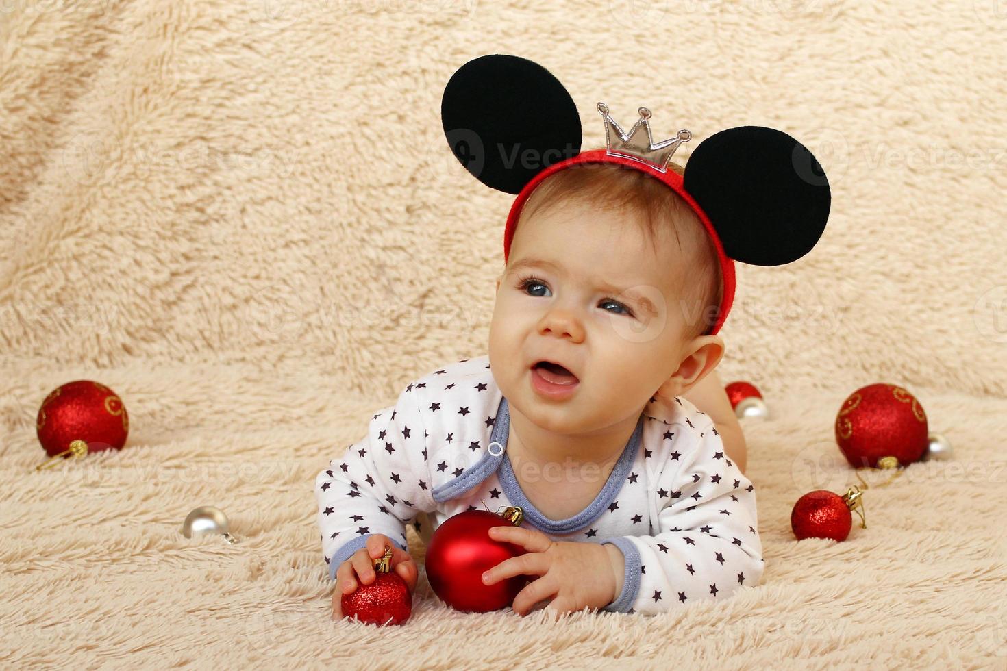 portrait d'une petite fille mignonne avec des oreilles de souris sur un plaid beige et des boules de noël brillantes rouges. photo