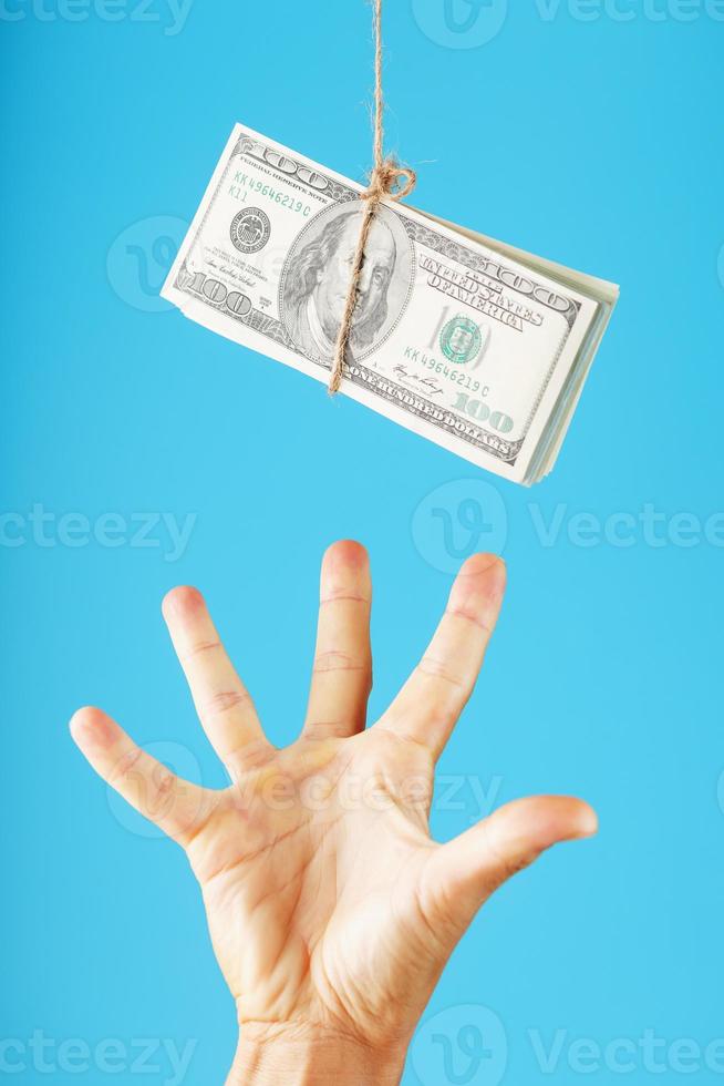 une main essaie de saisir une liasse d'argent sur une corde sur fond bleu. des billets d'un dollar sont suspendus à une corde. photo
