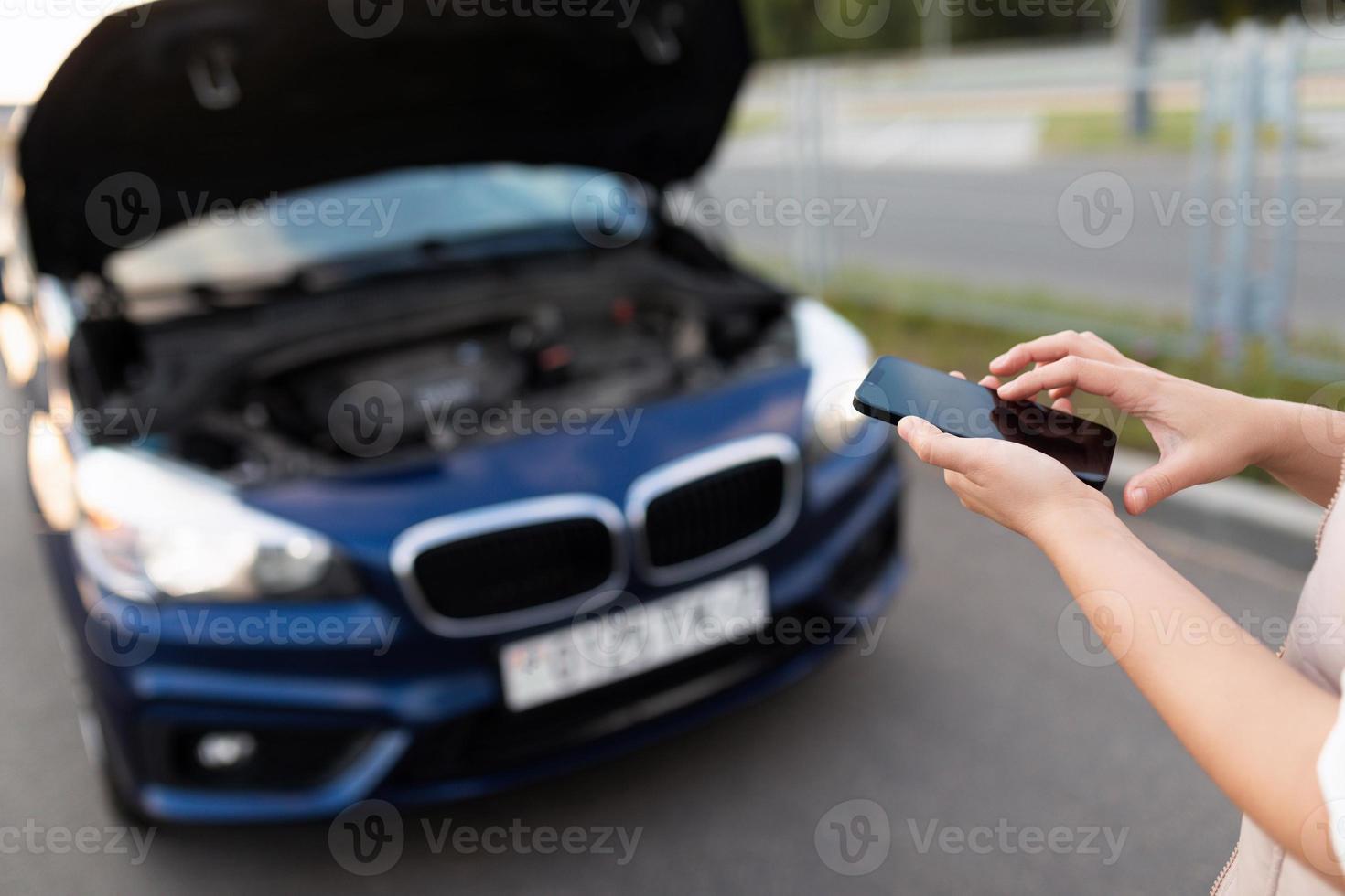 netteté sur un téléphone ekeran, une femme compose un numéro de dépanneuse ou d'assistance technique sur un téléphone portable à côté d'une voiture avec un capot ouvert, photo avec profondeur de champ