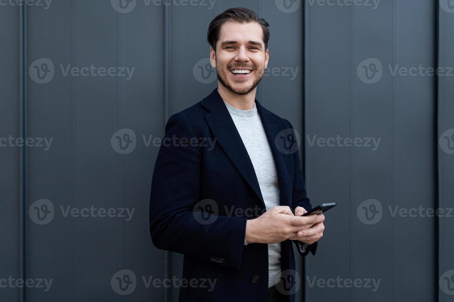 portrait d'un homme d'affaires d'âge moyen fort utilisant un téléphone portable et un large sourire contre un mur gris photo