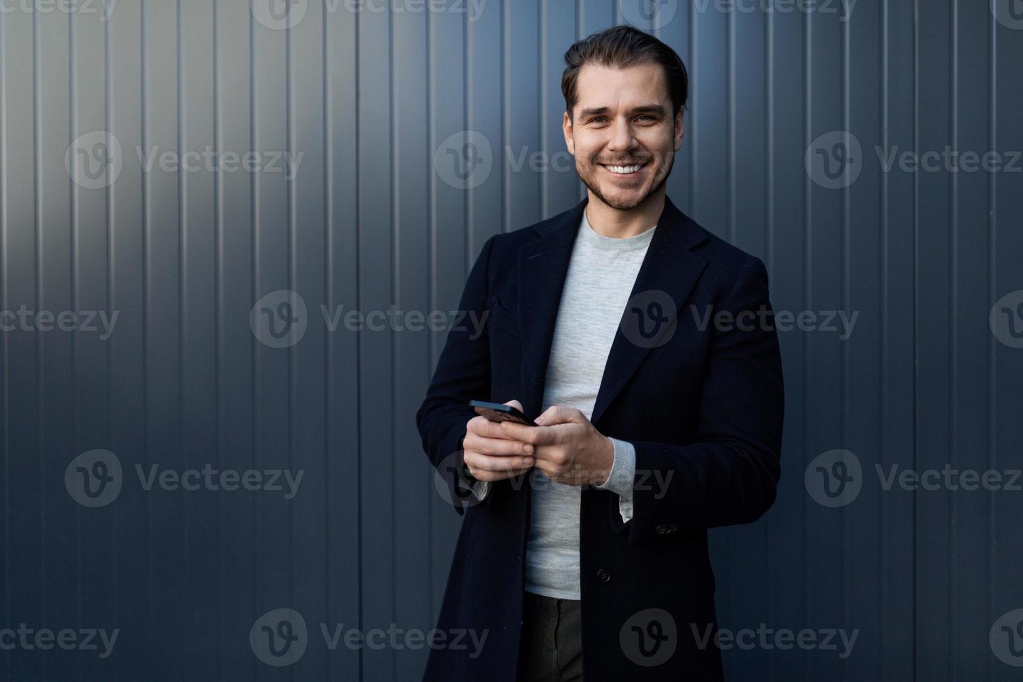 jeune ingénieur logiciel prospère avec un large sourire sur son visage et un téléphone portable dans ses mains contre un mur gris photo