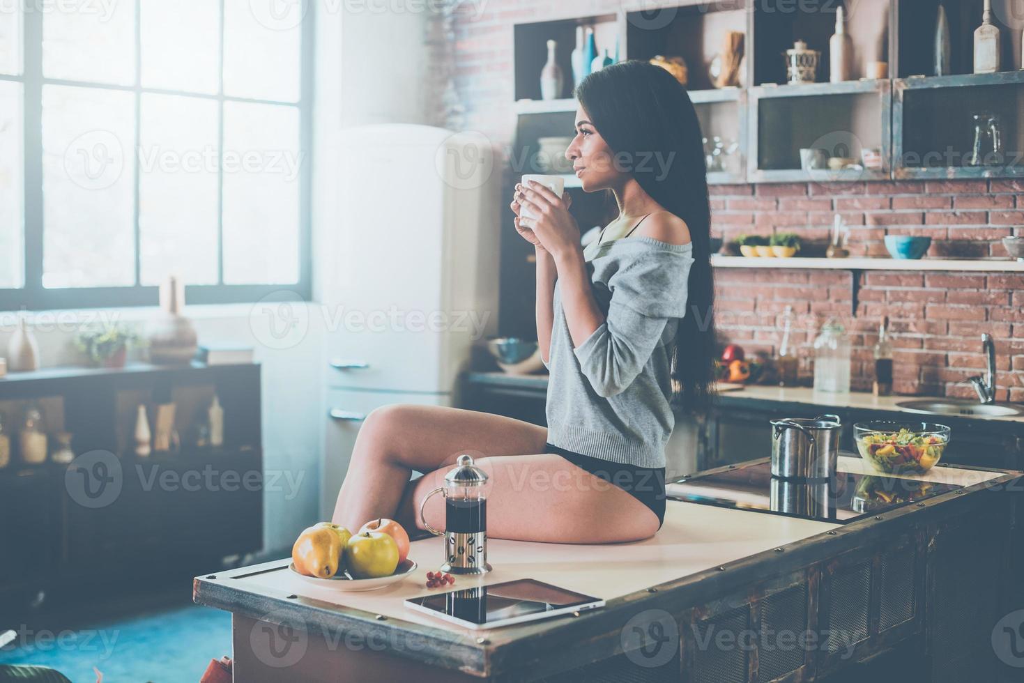 boire du café frais. belle jeune femme métisse tenant une tasse de café en détournant les yeux tout en étant assis sur le bureau de la cuisine photo