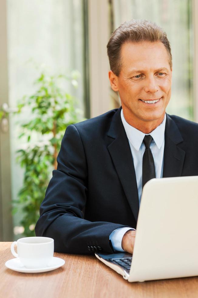 travailler avec plaisir. joyeux homme mûr en tenues de soirée travaillant sur un ordinateur portable et souriant tout en étant assis à la table à l'extérieur photo
