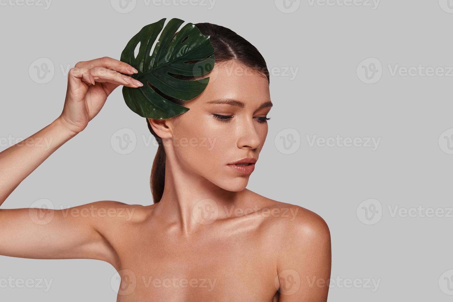 suprêmement sensuel. séduisante jeune femme regardant loin et couvrant de feuilles en se tenant debout sur fond gris photo