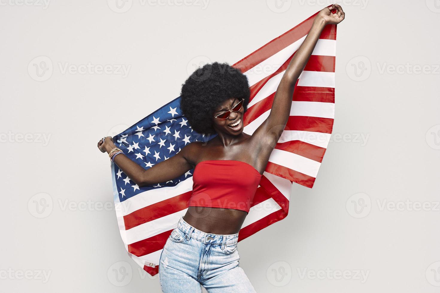 heureuse jeune femme africaine portant le drapeau américain et souriant en se tenant debout sur fond gris photo