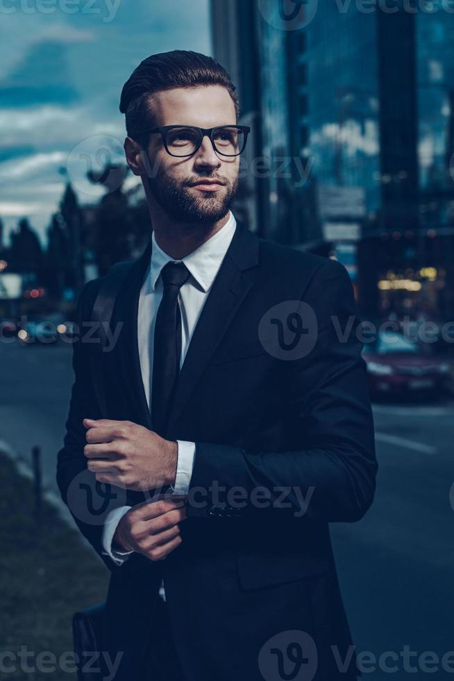 homme d'affaires moderne. jeune homme confiant en costume complet ajustant sa manche et regardant ailleurs tout en se tenant à l'extérieur avec le paysage urbain en arrière-plan photo