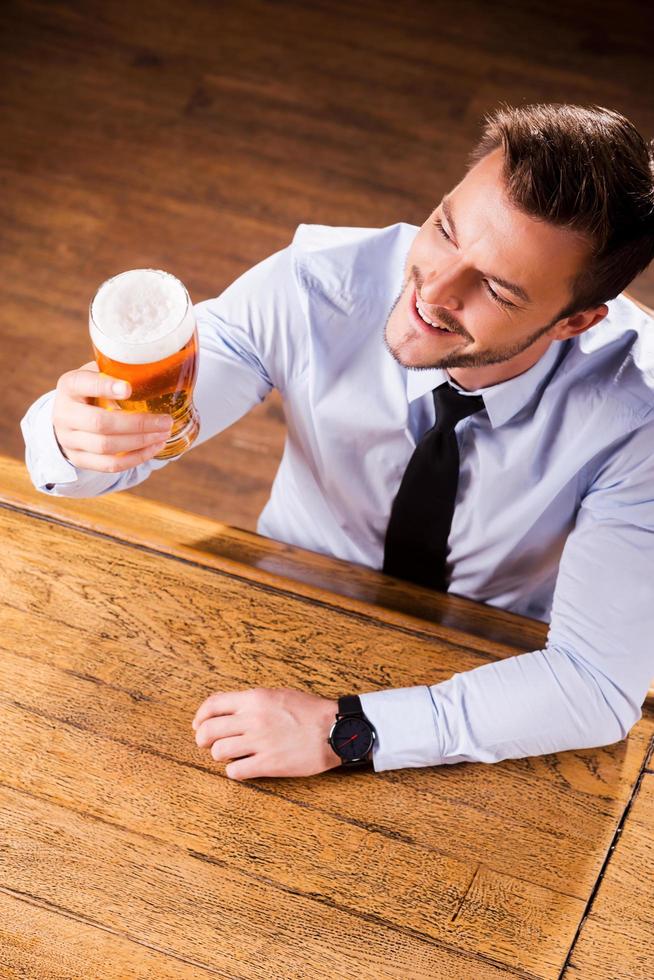 profiter de la meilleure bière de la ville. vue de dessus du beau jeune homme en chemise et cravate examinant le verre avec de la bière et souriant alors qu'il était assis au comptoir du bar photo