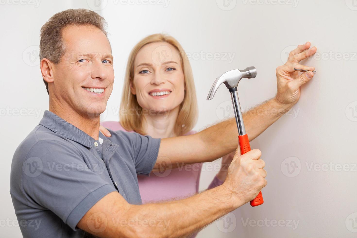 réparer la maison ensemble. joyeux homme aux cheveux gris martelant un clou et souriant tandis que sa femme se tenant près de lui photo