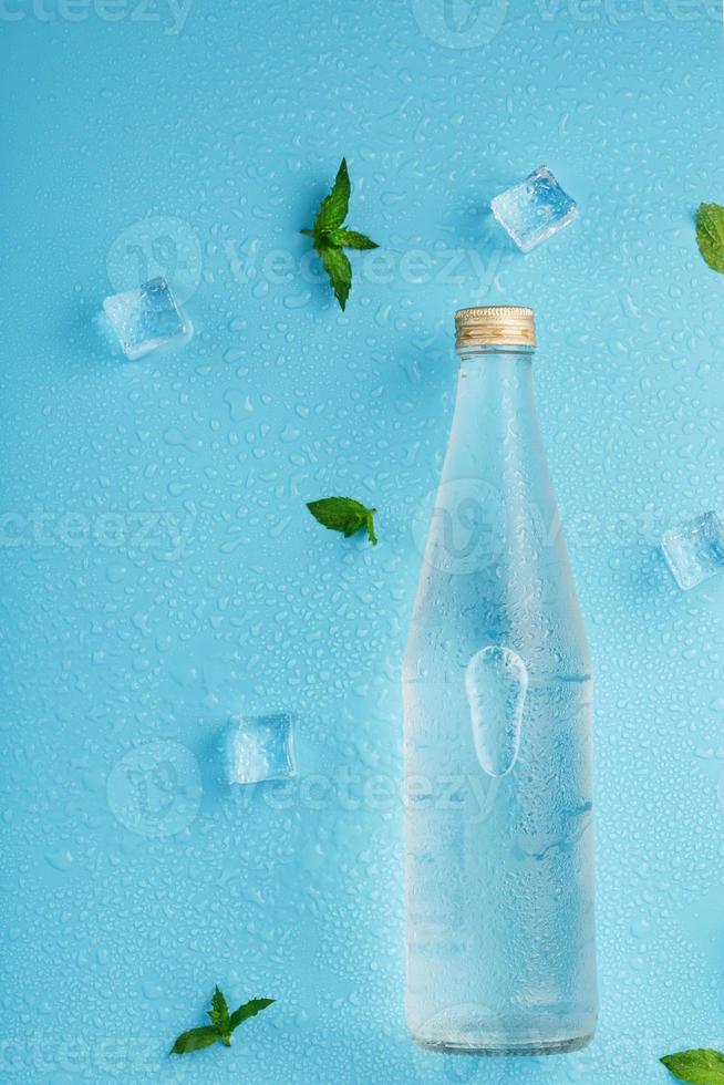 bouteille avec une boisson glacée, des glaçons, des gouttes et des feuilles de menthe sur fond bleu. photo