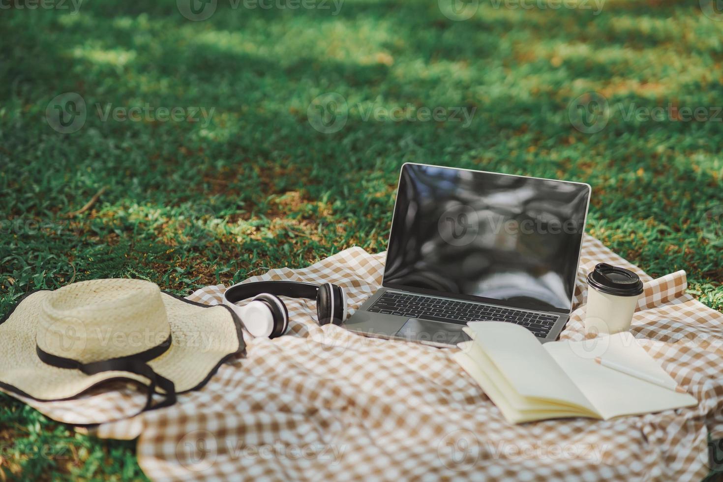 travaillez sur un ordinateur portable sur une couverture de pique-nique sur la pelouse du parc - à côté d'un casque, d'un cahier, d'un stylo, d'un chapeau et d'une tasse de café chaud. concept de travail indépendant. vue de dessus, mise à plat. forme de travail n'importe où concept. photo