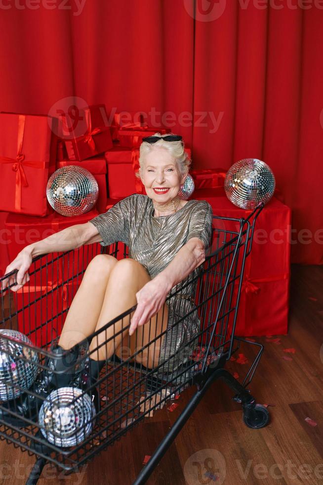 Senior élégante femme aux cheveux gris en lunettes de soleil et robe argentée assise dans le chariot de supermarché à la fête, buvant du vin sur fond de rideaux rouges. fête, disco, célébration, concept d'âge senior photo