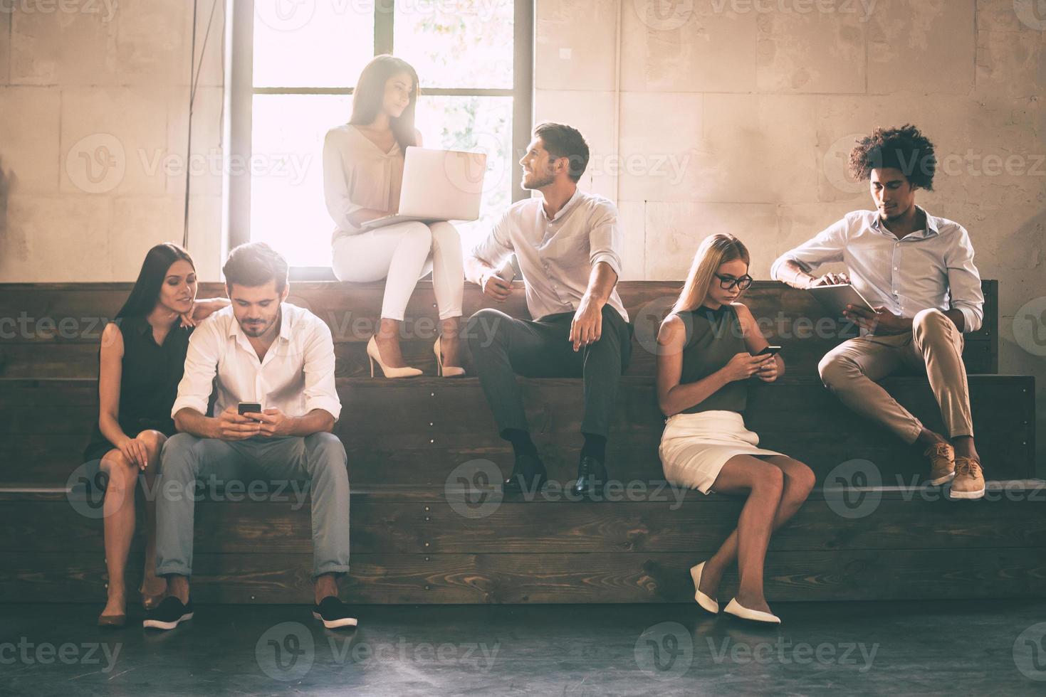 la vie des étudiants. groupe de jeunes joyeux communiquant tout en tenant différents gadgets et assis les uns à côté des autres sur les marches photo