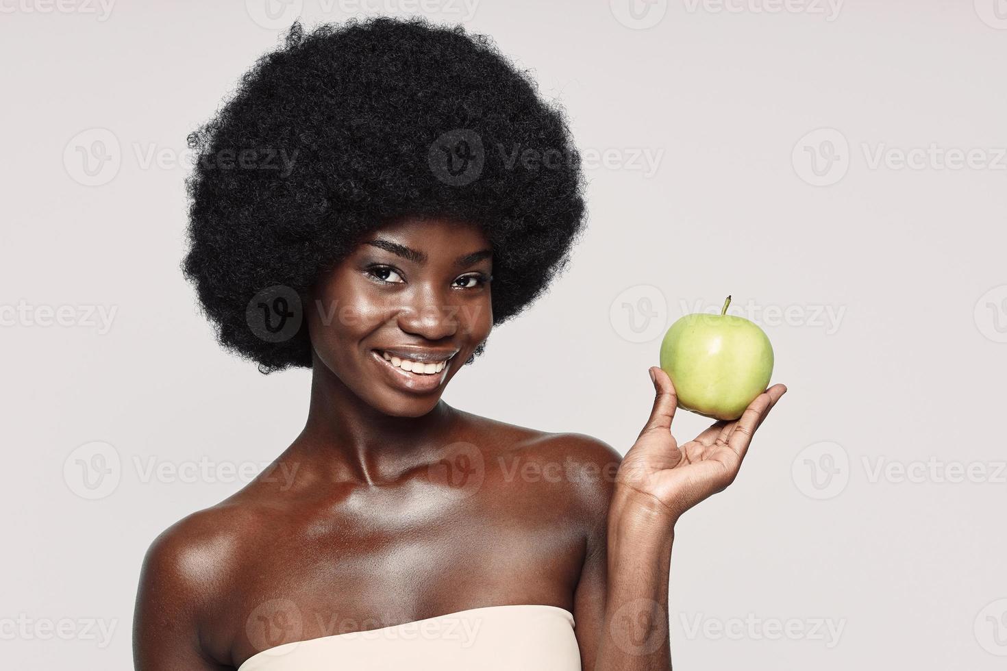 portrait de la belle jeune femme africaine tenant une pomme verte et souriant debout sur fond gris photo