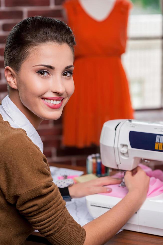 elle est la meilleure de l'industrie de la mode. vue arrière d'une créatrice de mode féminine joyeuse travaillant sur une machine à coudre et regardant par-dessus l'épaule tout en étant assise sur son lieu de travail photo