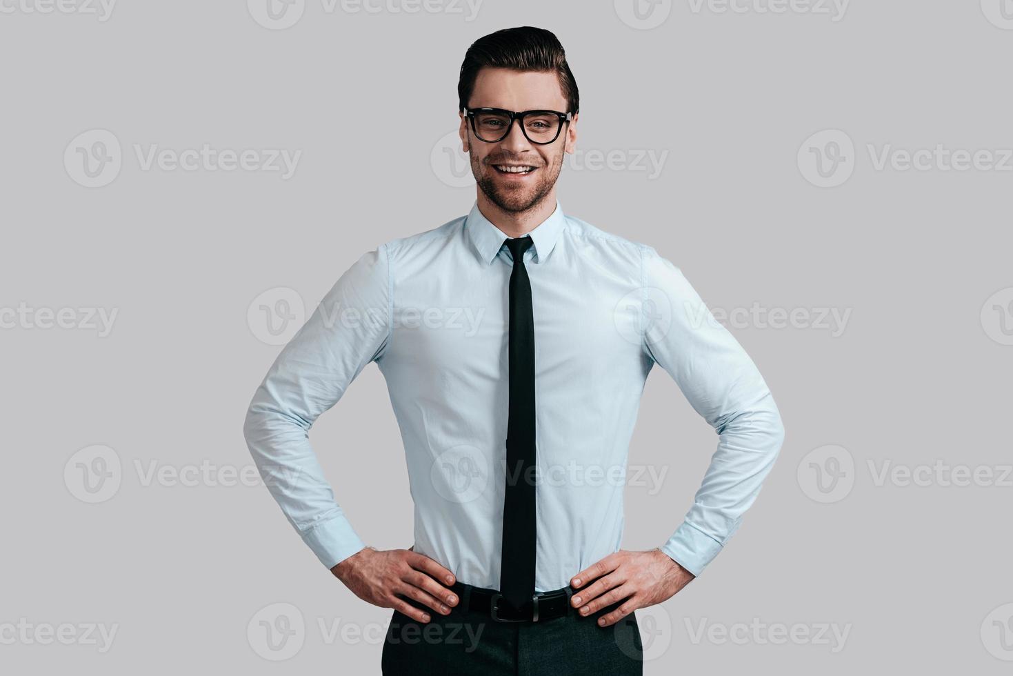 jeune et prospère. beau jeune homme à lunettes en gardant les mains sur la hanche et en regardant la caméra en se tenant debout sur fond gris photo