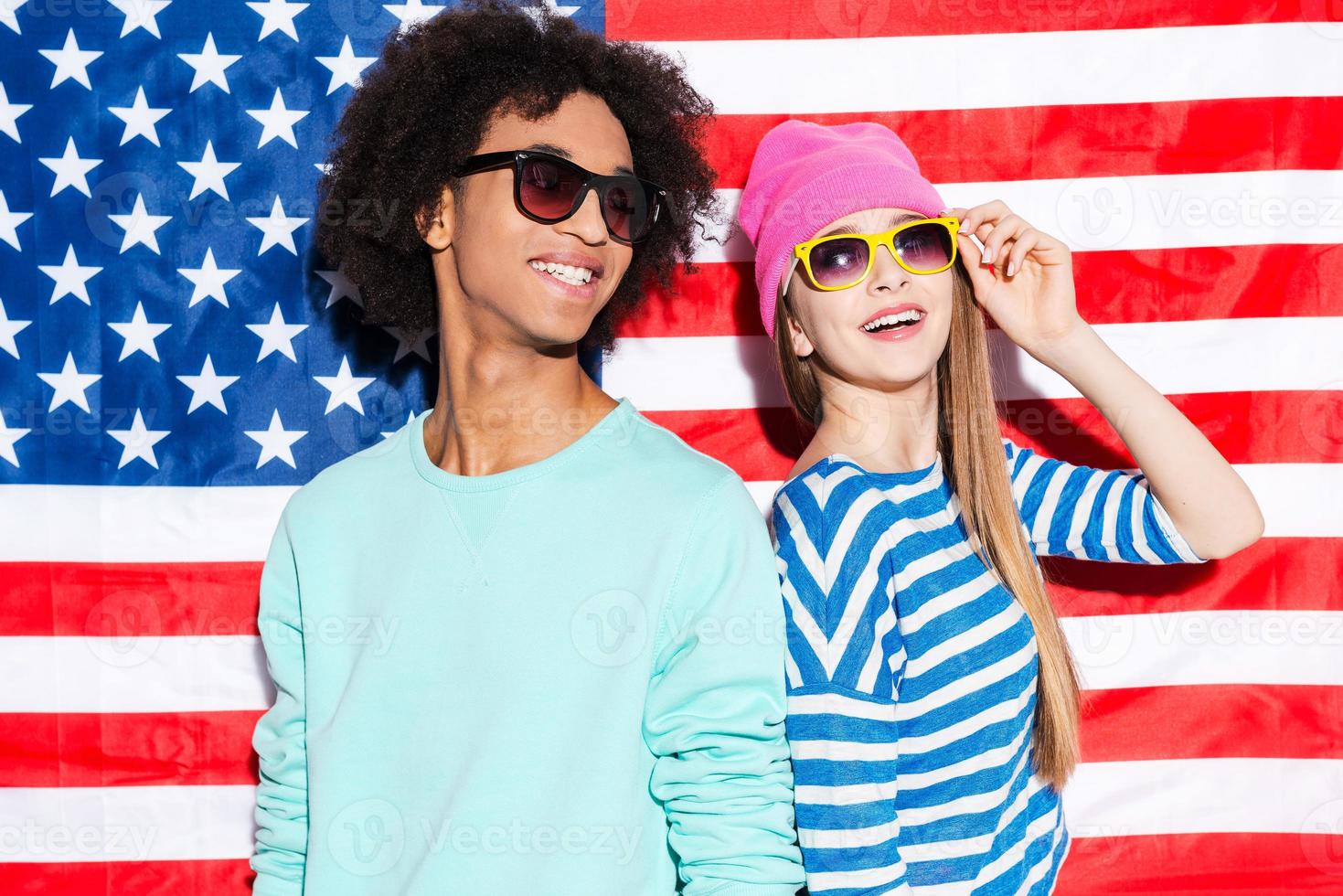 rêves américains. funky jeune couple portant des lunettes de soleil et souriant debout contre le drapeau américain photo