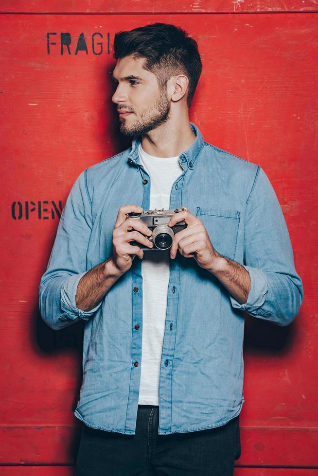 beau photographe. beau jeune homme à lunettes de soleil en gardant les bras croisés et en regardant la caméra en se tenant debout sur fond rouge photo