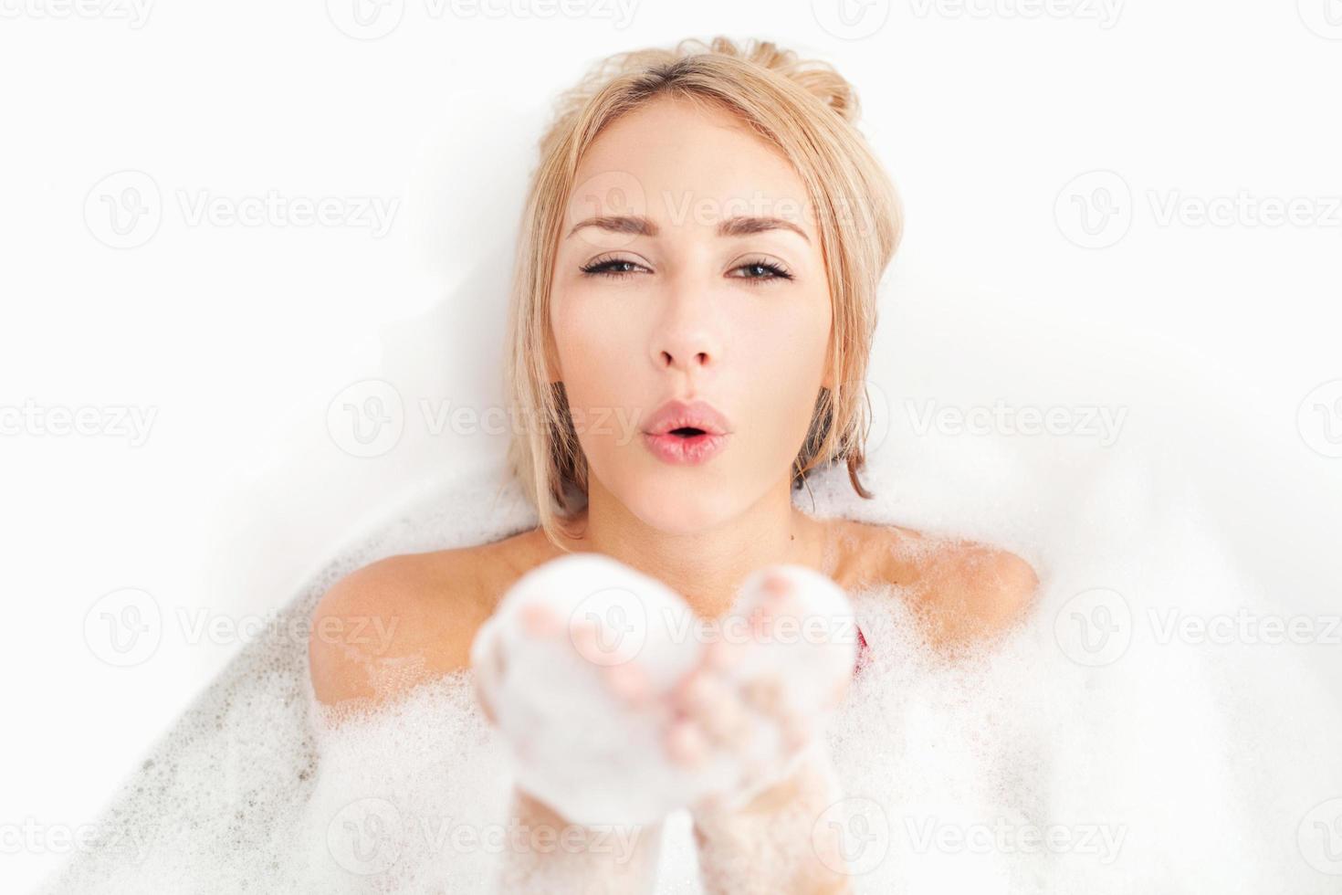 bain de beauté. jeune femme ludique allongée dans une baignoire pleine de mousse savonneuse et soufflant un baiser d'air photo