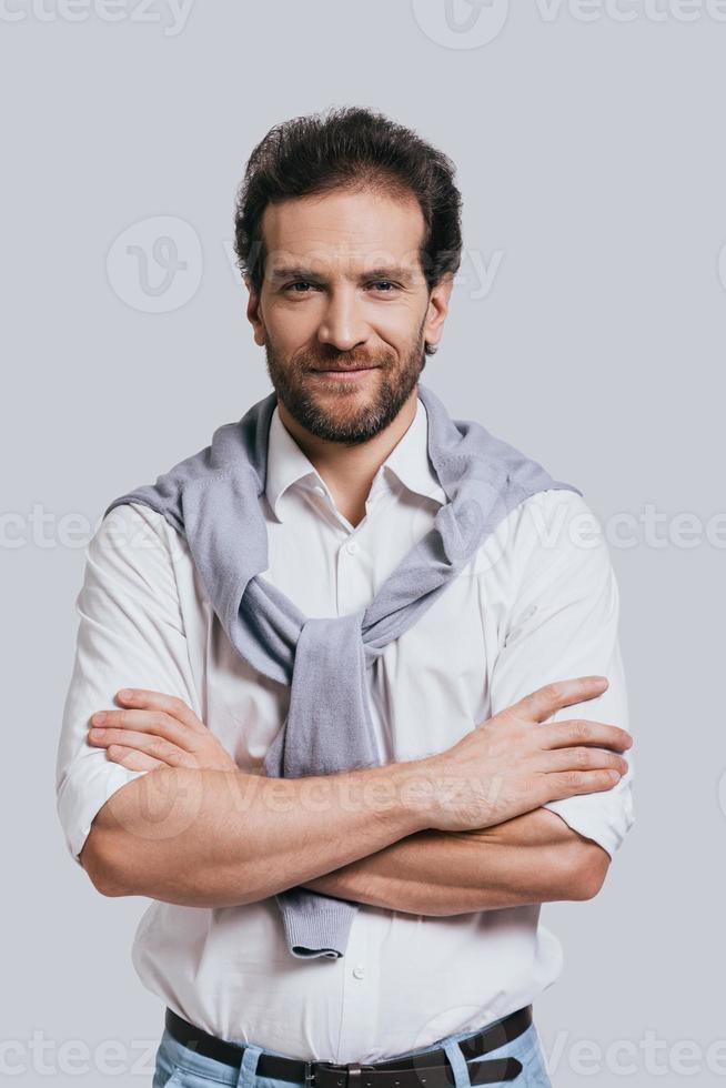 confiance et style. beau jeune homme gardant les bras croisés et regardant la caméra en se tenant debout sur fond gris photo