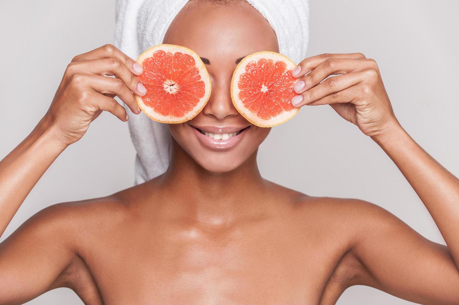 station thermale naturelle. belle jeune femme torse nu afro-américaine tenant des morceaux d'orange devant ses yeux en se tenant debout isolé sur fond gris photo