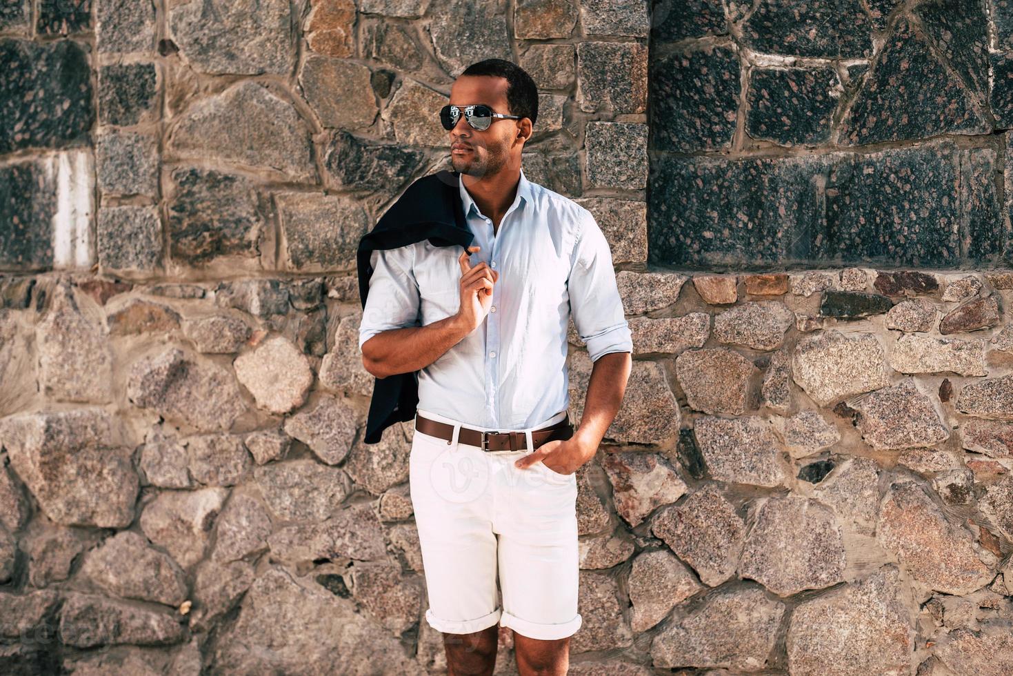 élégant beau. beau jeune homme africain en vêtements décontractés intelligents portant une veste sur l'épaule et regardant loin debout contre le mur de pierre à l'extérieur photo