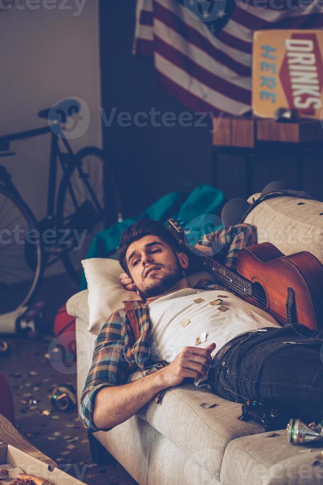 jeune bel homme regardant loin en position couchée sur le canapé dans une pièce en désordre après la fête photo