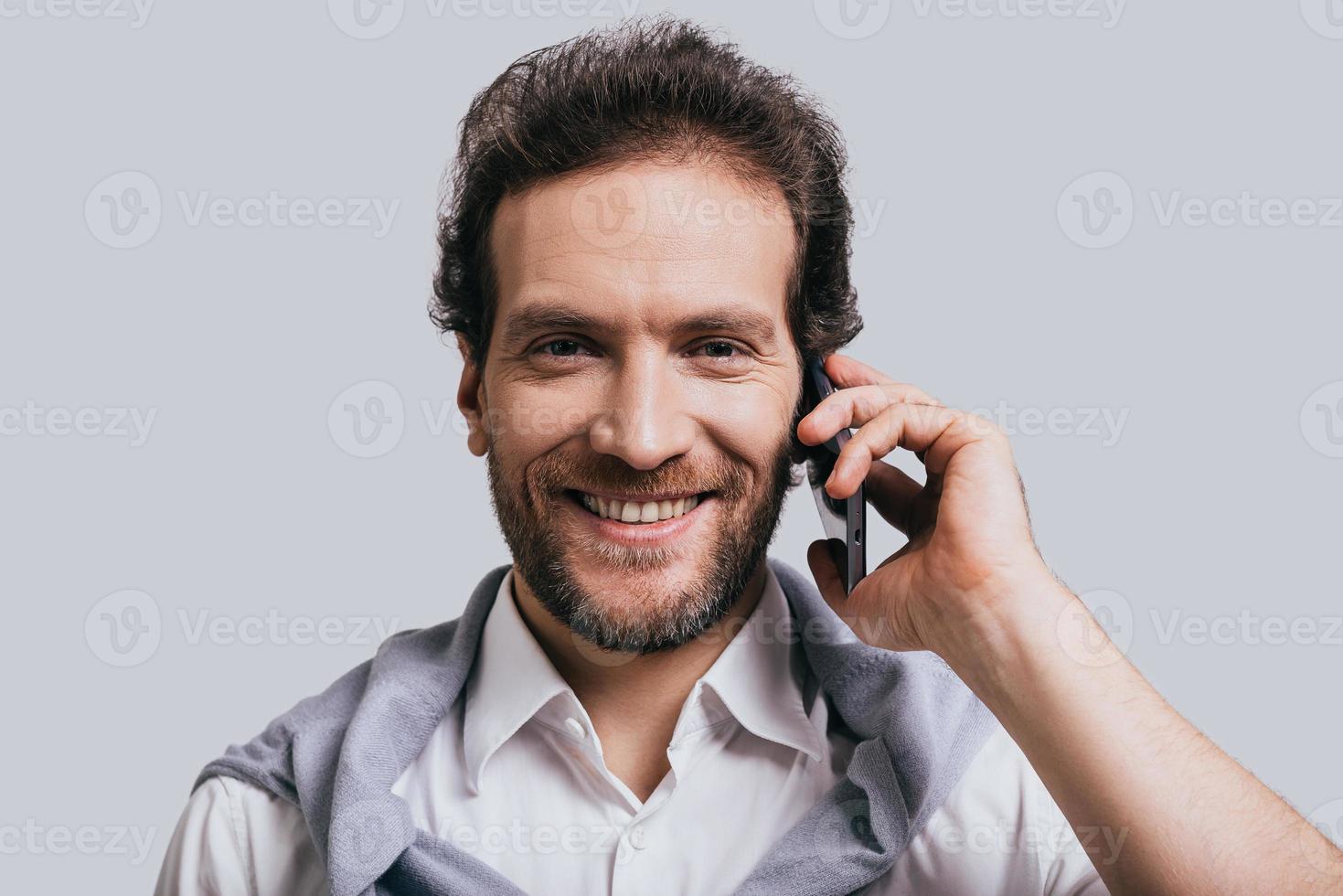 conversation agréable. beau jeune homme en vêtements décontractés intelligents parlant au téléphone et regardant la caméra en se tenant debout sur fond gris photo